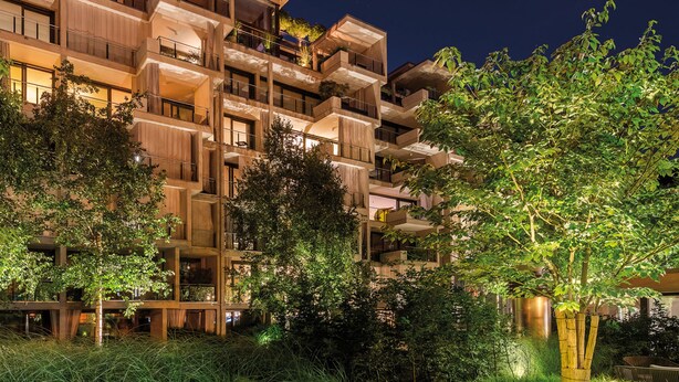 A modern outdoor lounge area with wicker furniture and a wooden deck, surrounded by lush greenery and a multi-story building in the background. - Hotel the YARD Berlin (Berlin)