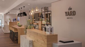 A reception area with a wooden desk, bar stools, and a modern chandelier. There are shelves with plants and a sign that reads "the YARD HOTEL". - Hotel the YARD Berlin (Berlin)