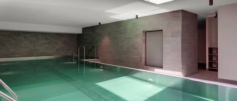 Piscina interior moderna com paredes de vidro transparente, escada de metal e claraboia.