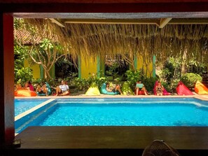 Un área de piscina con techo de paja, rodeada de follaje y coloridos puffs.