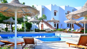 Un área de piscina con sillones de descanso y sombrillas de paja, una escalera y un edificio blanco con ventanas arqueadas.