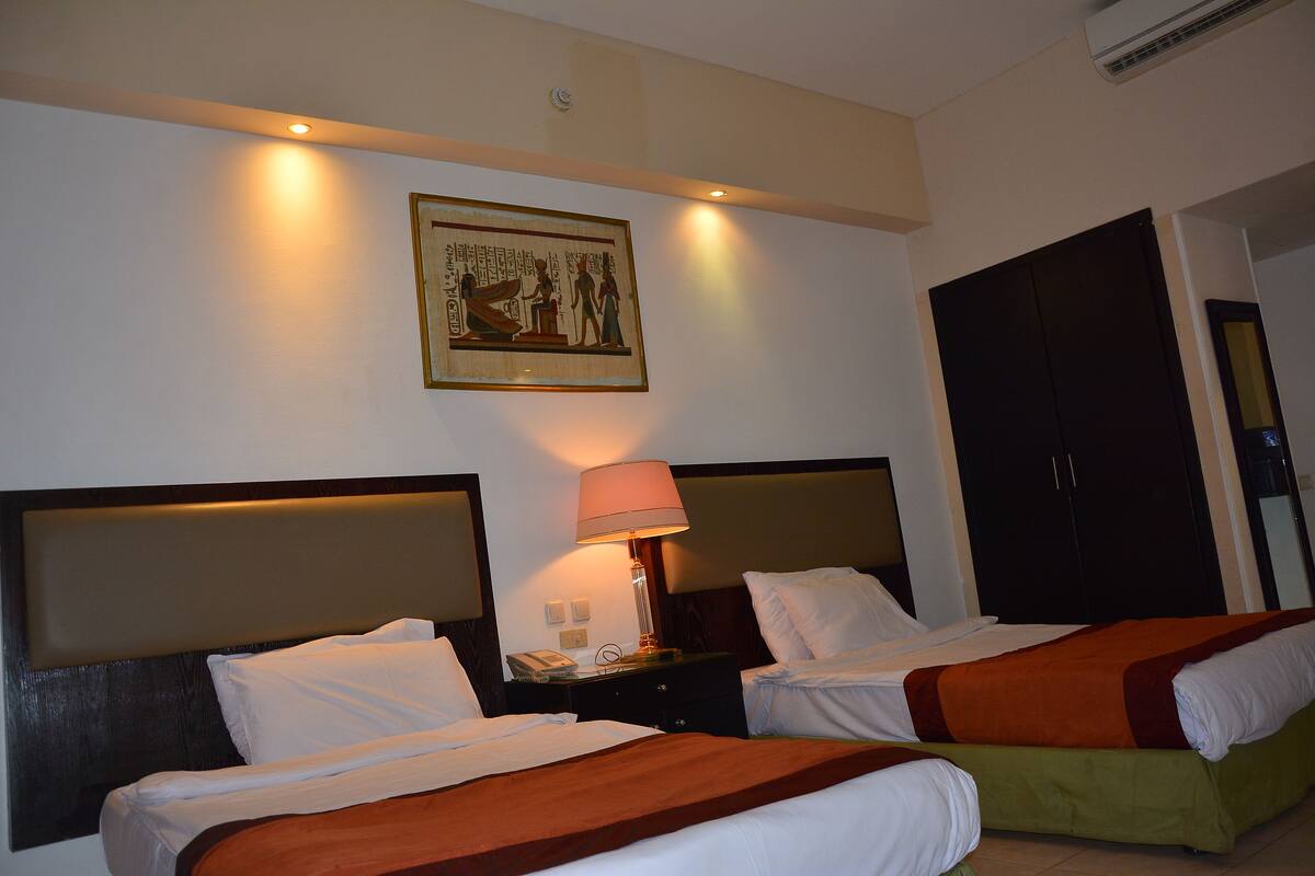 Habitación de hotel con dos camas, una mesita de noche con lámpara y un cuadro colgado en la pared.