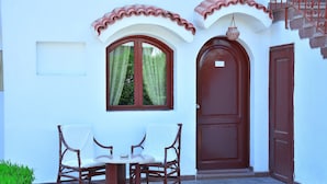 Un patio con dos sillas de mimbre, una mesita redonda, una puerta marrón y una ventana con cortina.