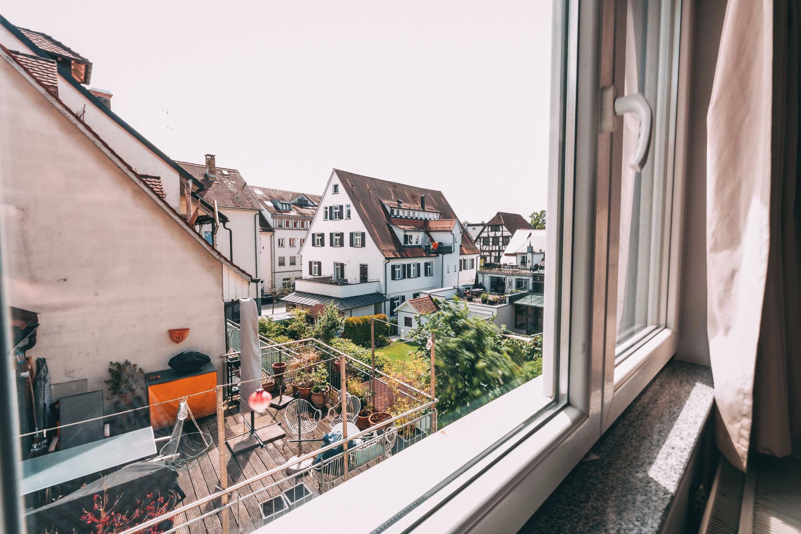 Une vue depuis une fenêtre donnant sur une rangée de bâtiments de style européen traditionnel, avec des balcons et des plantes en pot visibles.