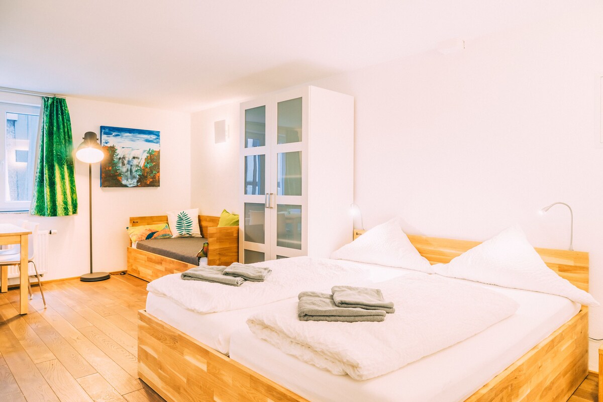 A hotel room with a wooden bed, a desk, a chair, a green curtain, and a painting on the wall.
