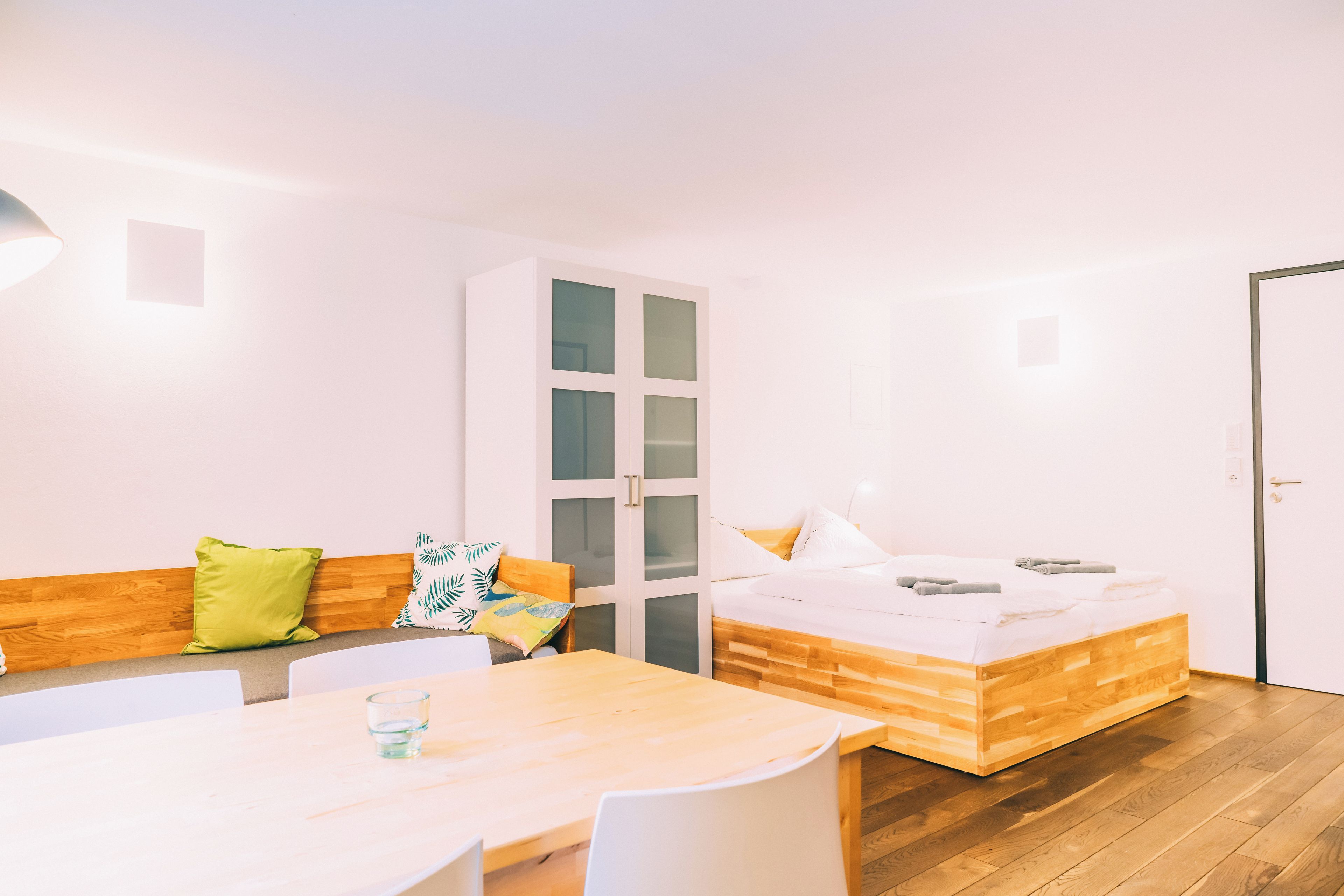 A modern room with a wooden bed, a white sofa with cushions, a dining table, and a glass of water.