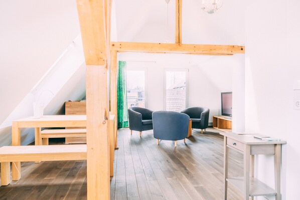 Un loft moderno con suelo de madera, una cama entrepiso, dos sillones oscuros, una pequeña mesa de madera y un televisor.