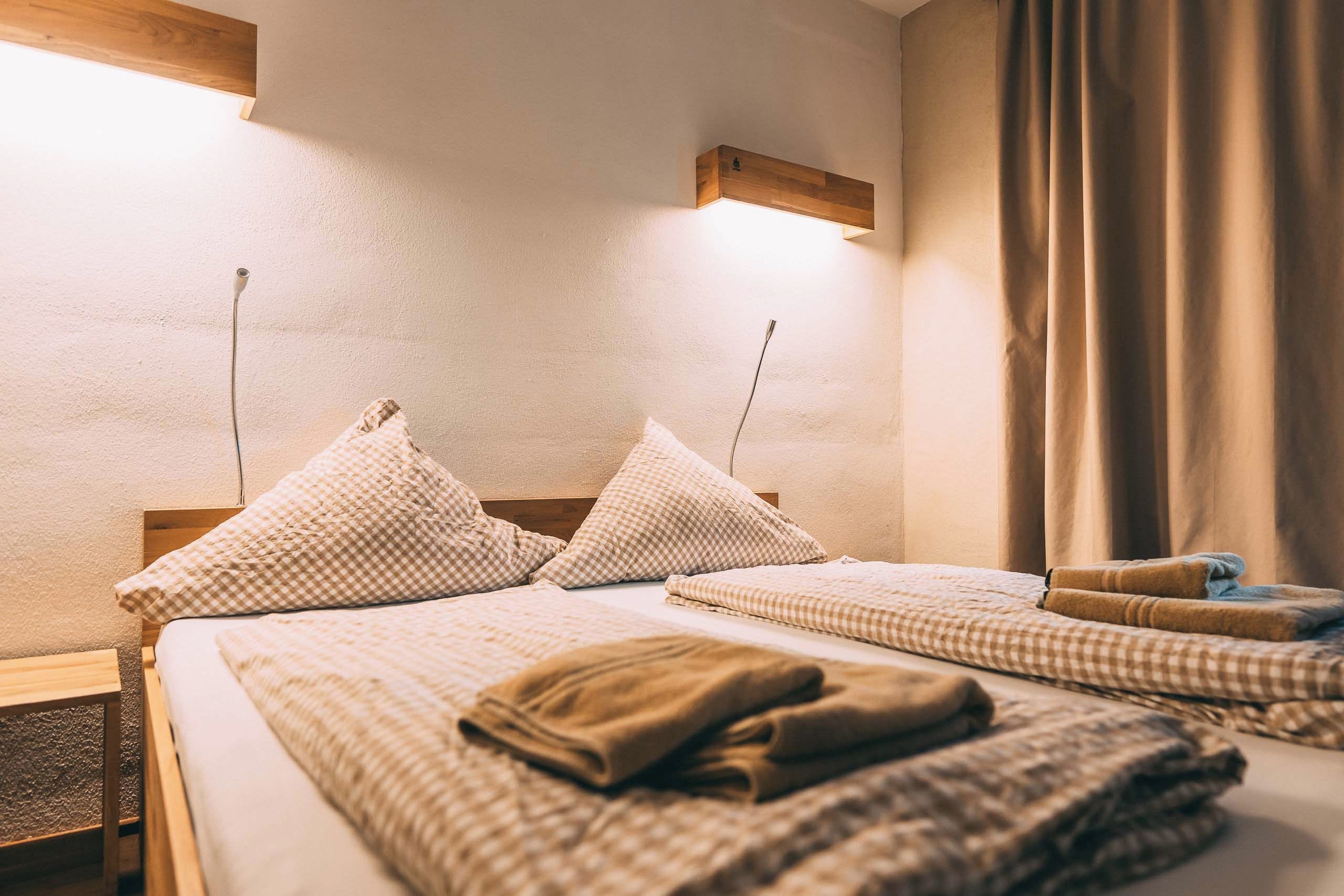 A hotel room with a bed, two pillows, a blanket, a wooden headboard, a wall-mounted light, and a window with curtains.