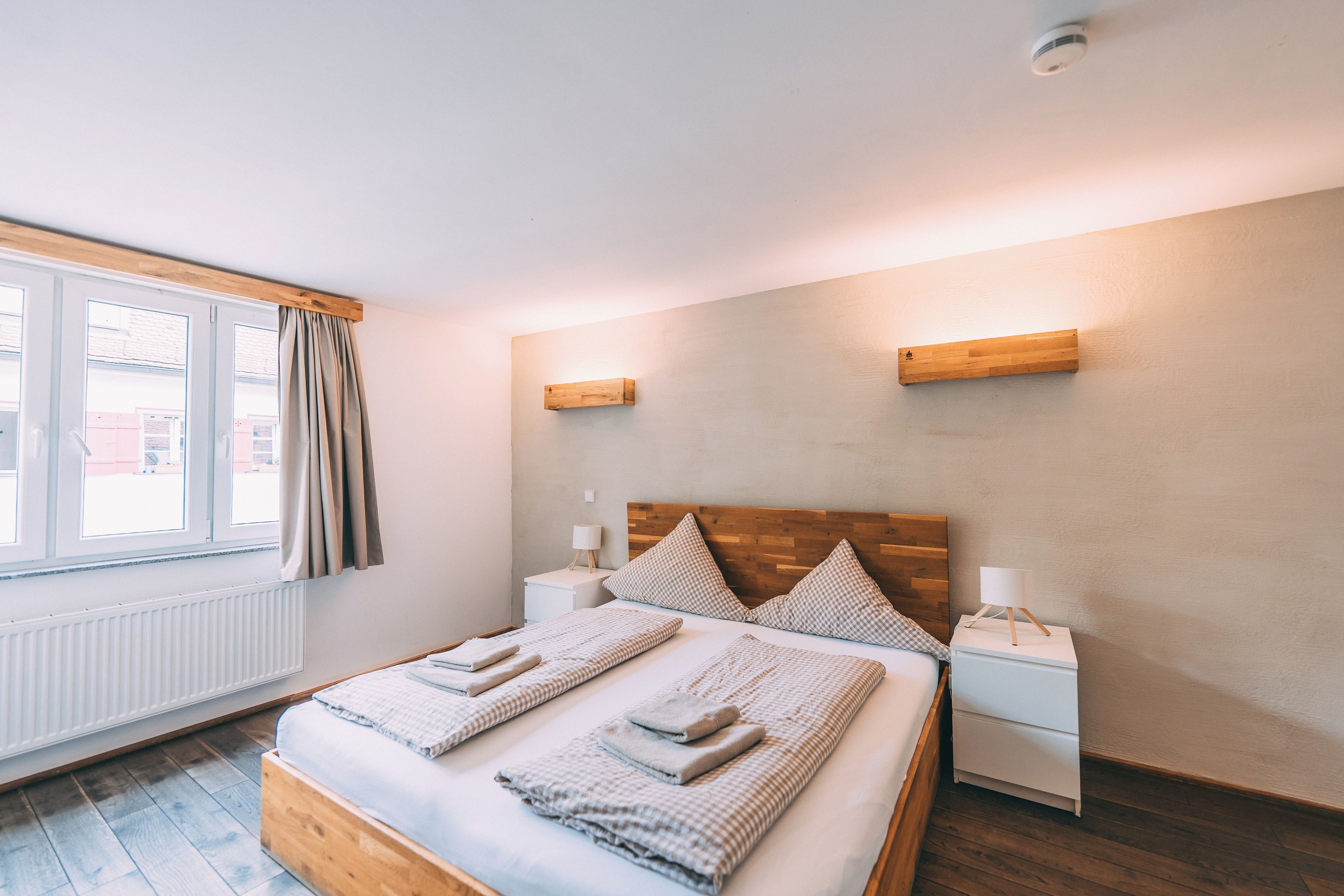 A bedroom with a wooden bed, two bedside tables, a window with curtains, and a wall-mounted light fixture.
