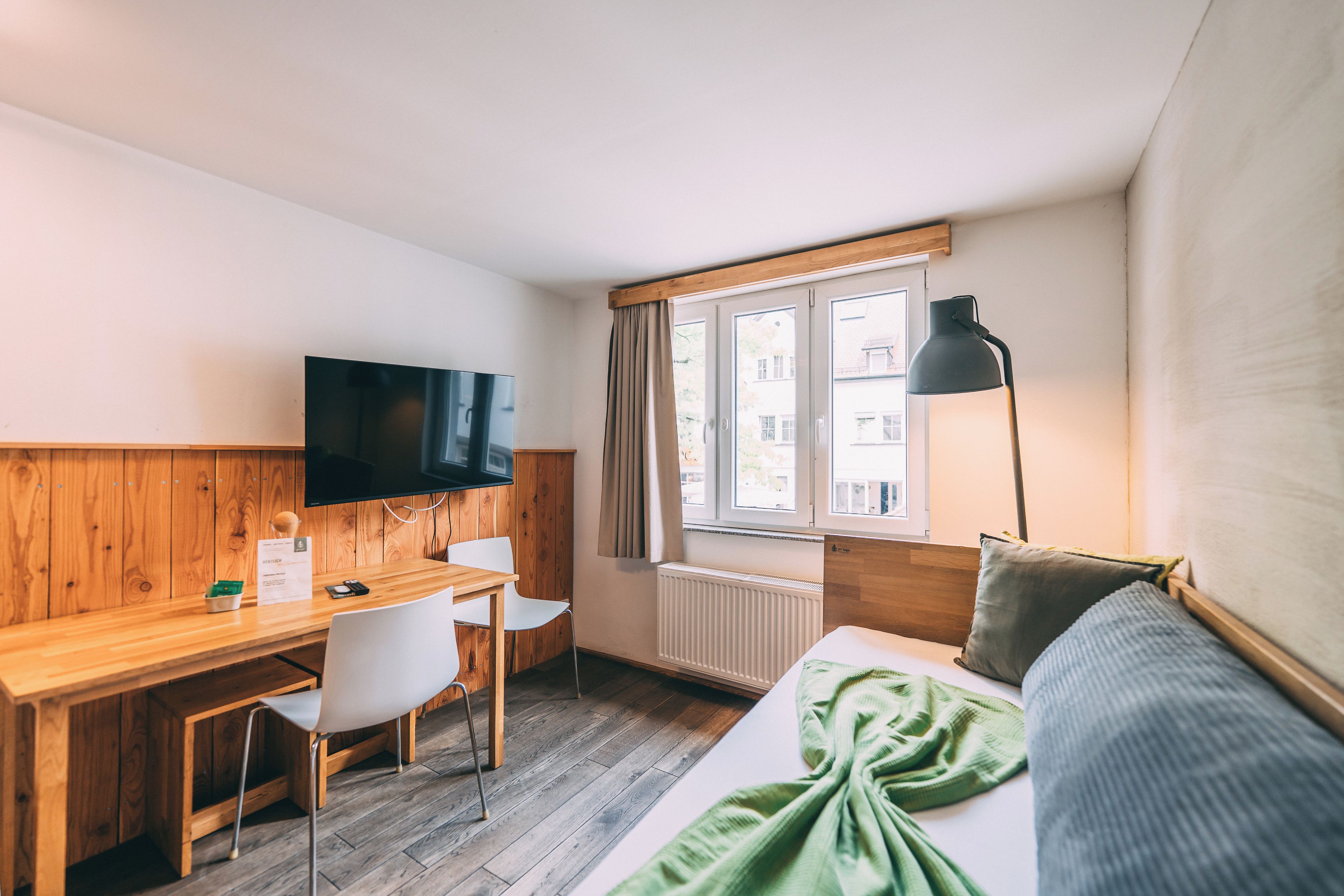Habitación de hotel con un escritorio de madera, un televisor, una ventana con cortinas, una cama con una manta verde y una lámpara de pie.