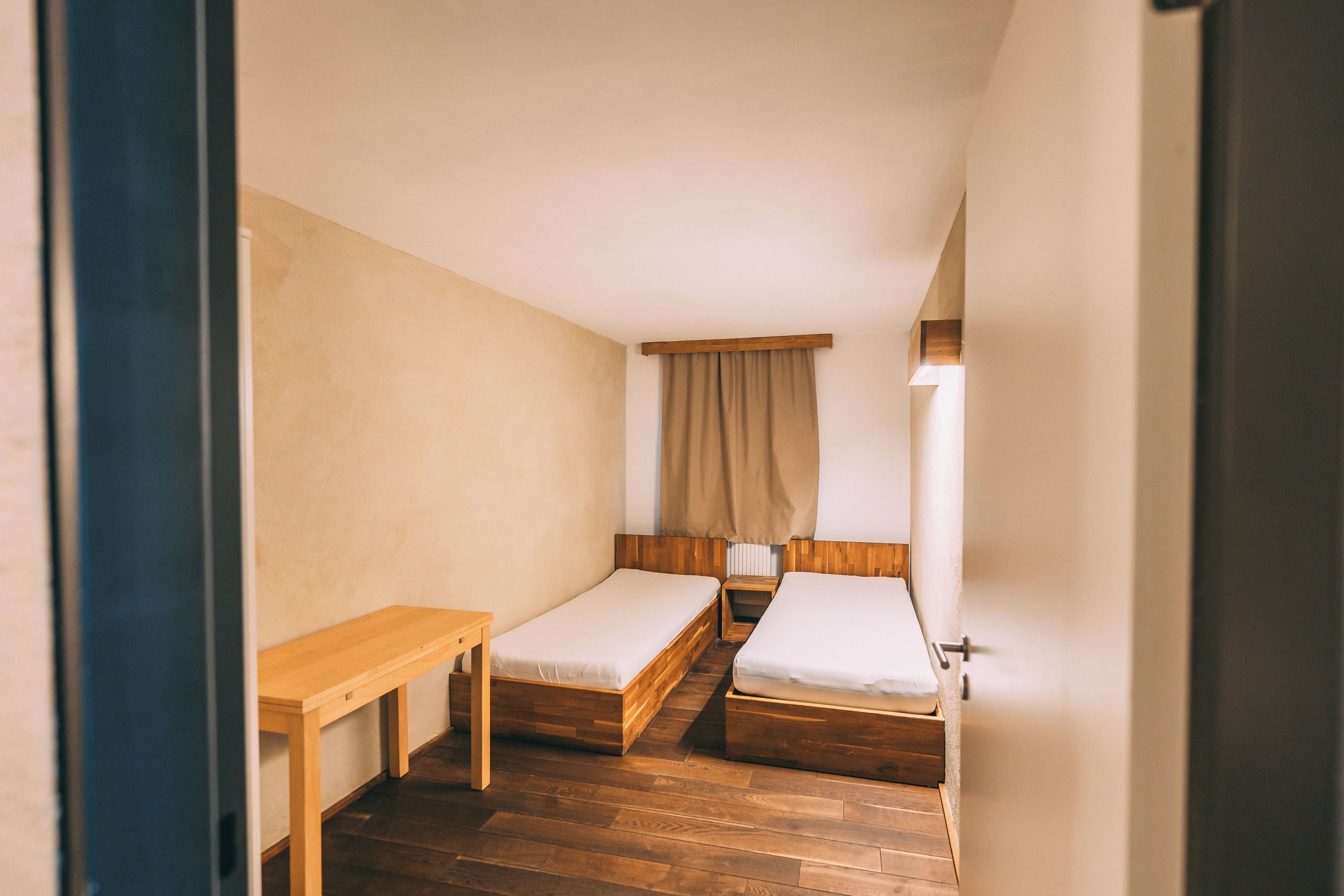 A small, compact room with two single beds, a wooden table, and a curtain.