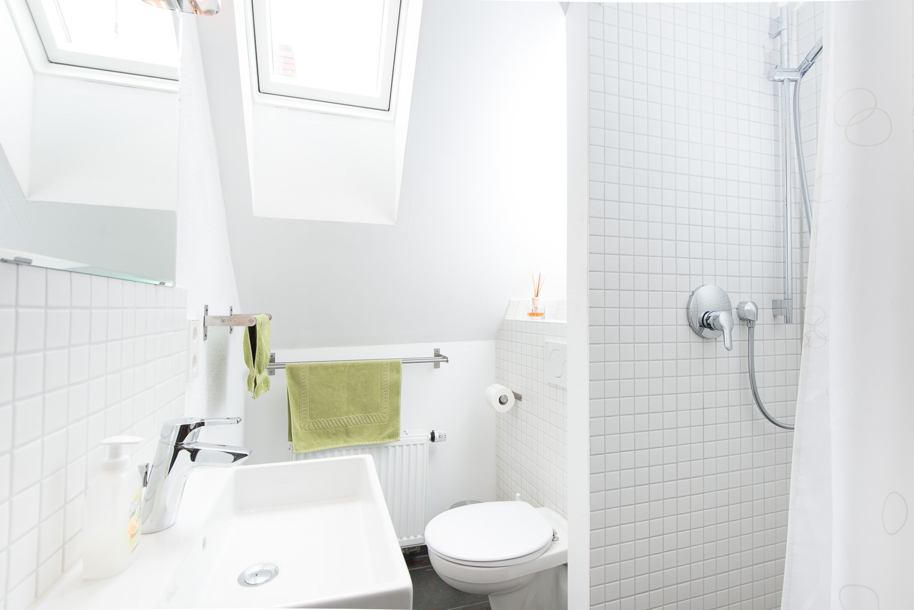 A modern bathroom with a white sink, toilet, and shower. There is a towel rack with a green towel, and a window above the sink.