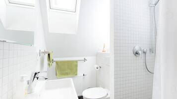 A modern bathroom with a white sink, toilet, and shower. There is a towel rack with a green towel, and a window above the sink.