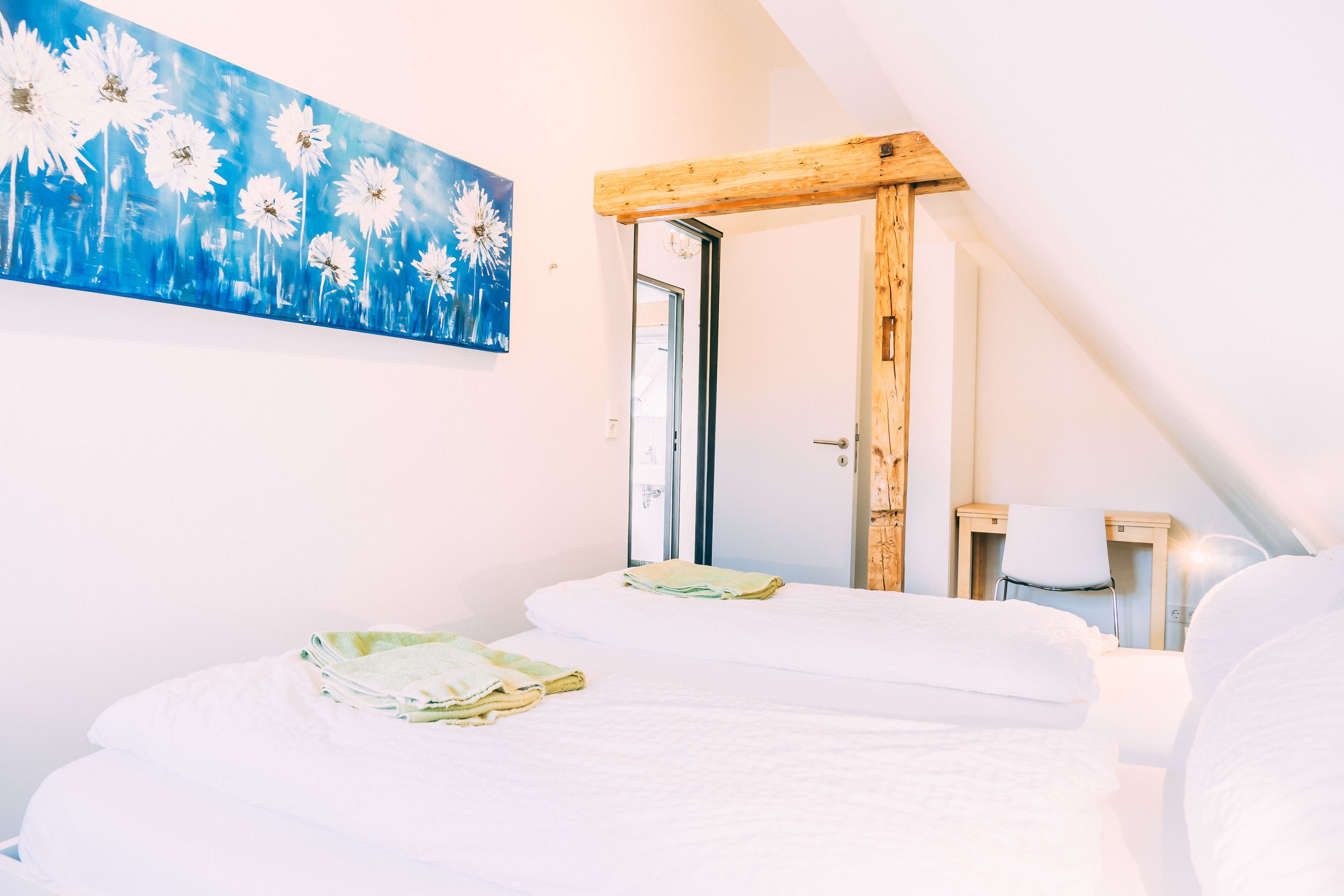 A bedroom with two beds, a wooden ceiling beam, a painting of daisies, and a view of the outdoors.