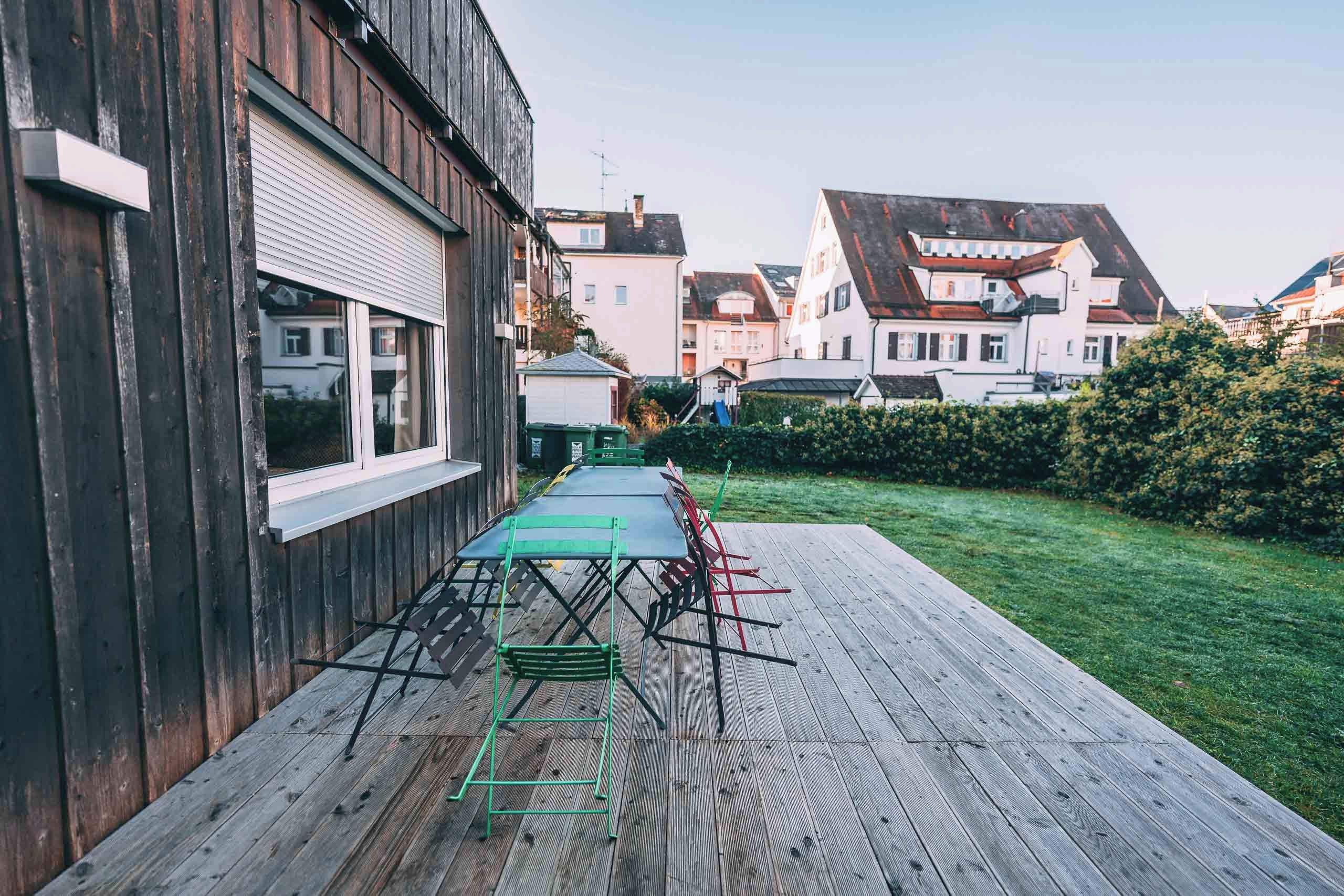 Une terrasse en bois avec une table et des chaises, une pelouse et des immeubles résidentiels en arrière-plan.