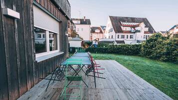 Una terraza de madera con una mesa y sillas, un césped y edificios residenciales al fondo.