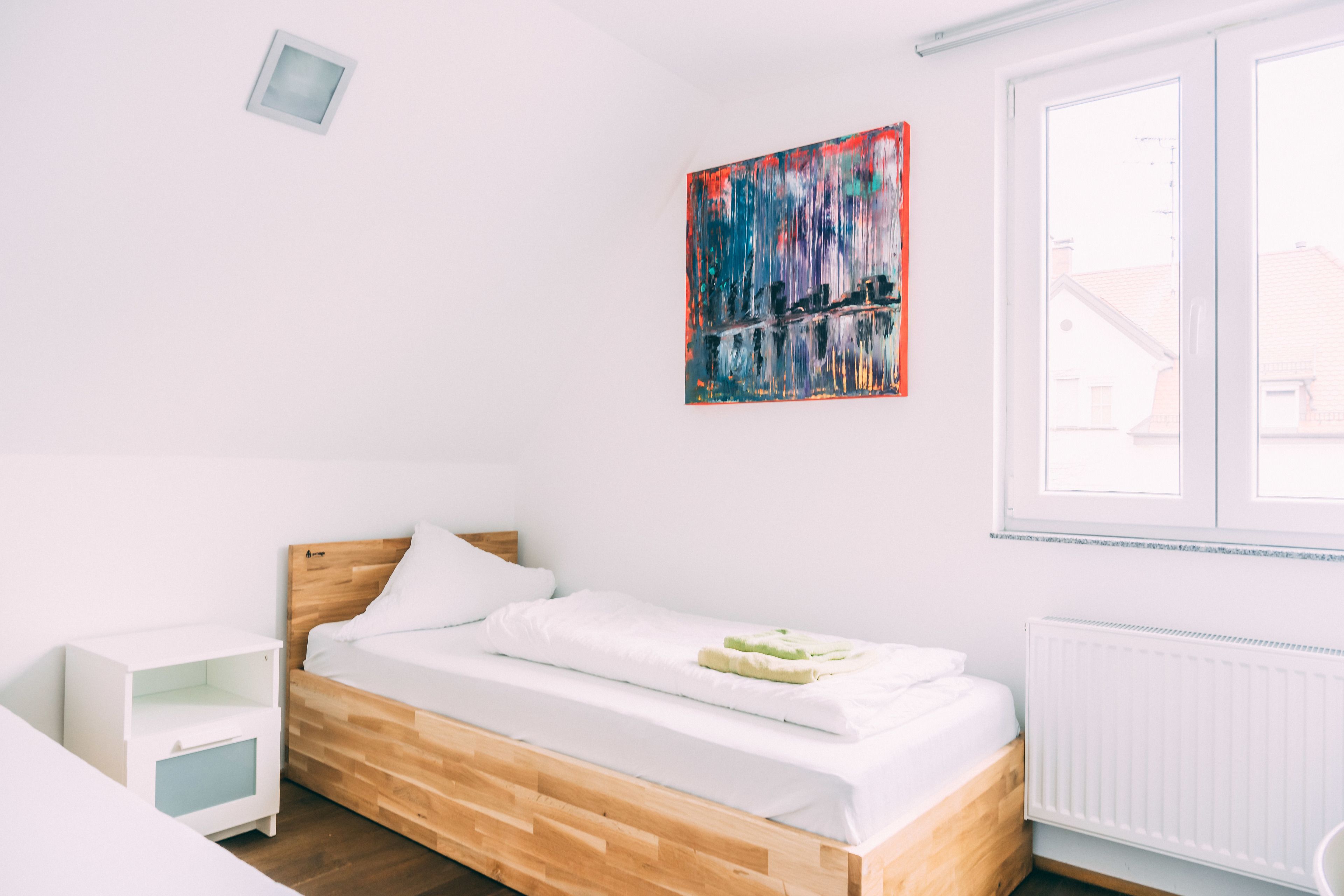 Een moderne slaapkamer met een houten bed, witte beddengoed, een klein nachtkastje en een kleurrijk abstract schilderij aan de muur.