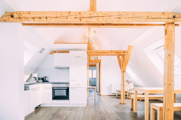 A modern kitchen with white cabinets, a built-in oven, and a coffee machine. There is a wooden dining table with chairs and a chandelier. - Stadthaus Gut Hügle (Ravensburg)
