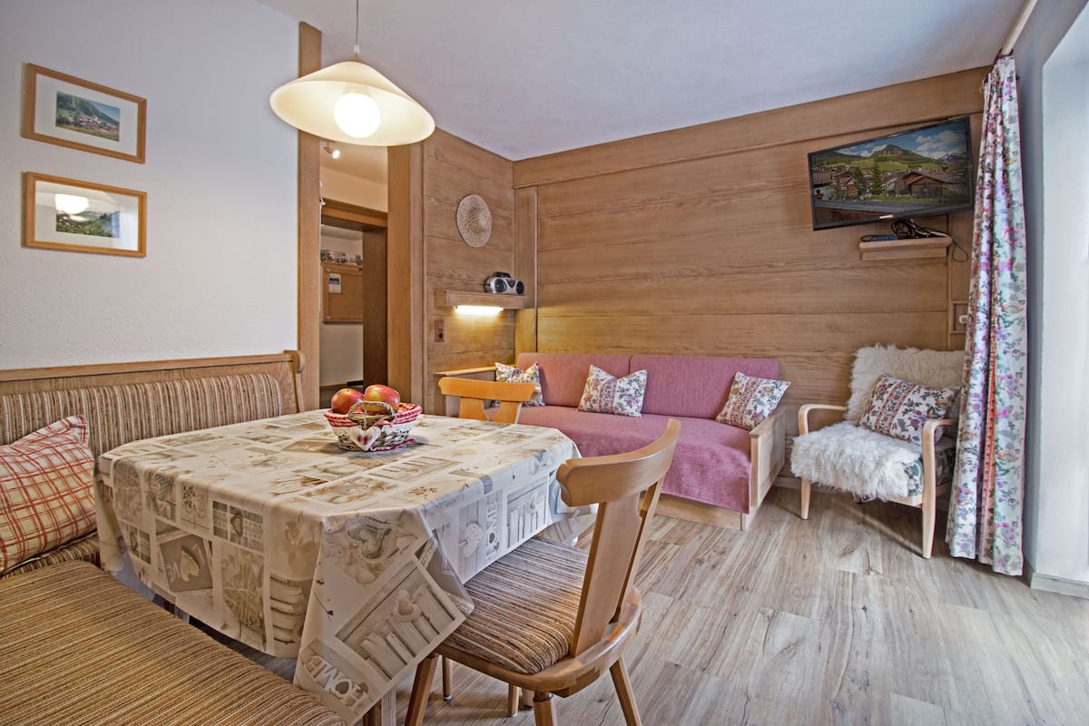 A cozy living room with a round table, a sofa, and a chair. There is a television mounted on the wall and a window with curtains.