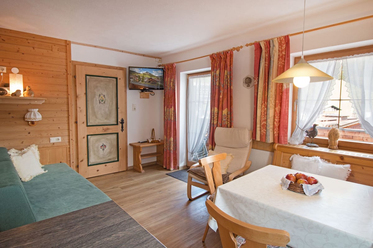 A cozy room with a wooden floor, a green sofa, a dining table with a basket of fruit, and a window with curtains.