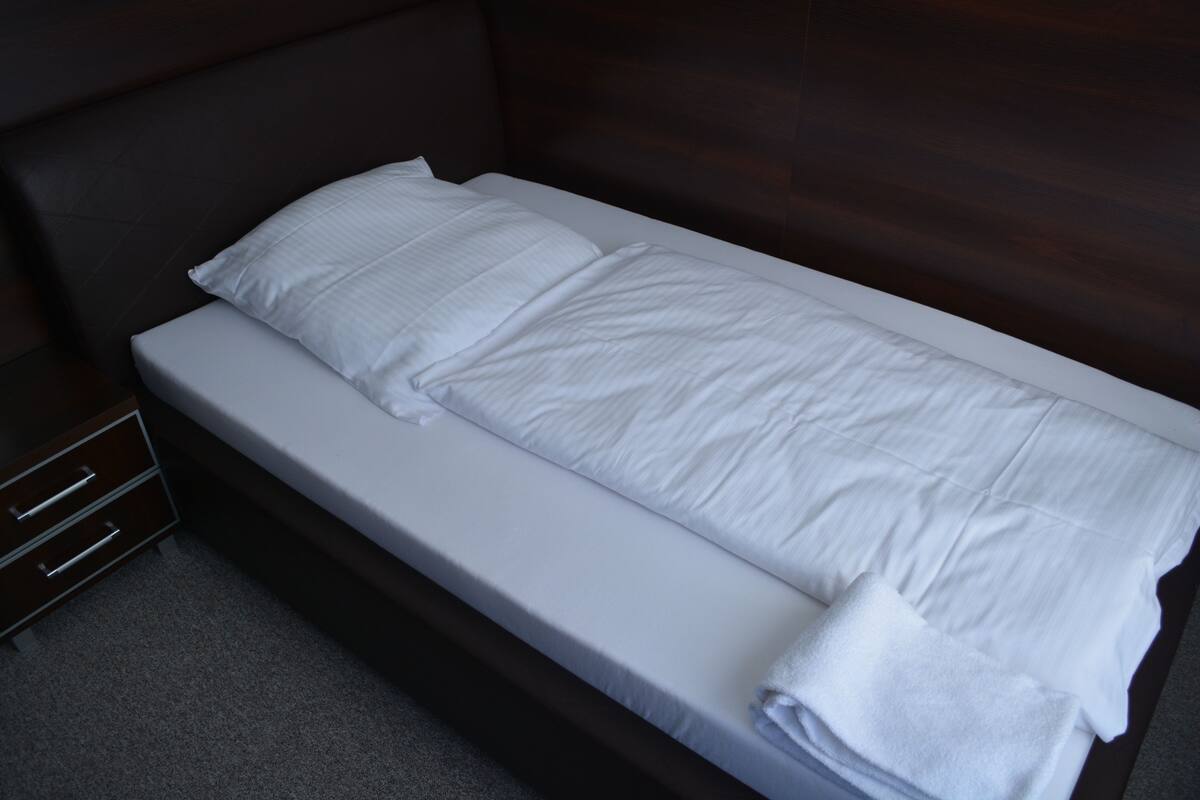 Una cama individual con ropa blanca, en una habitación con cabecera de madera oscura y una pequeña mesita de noche.
