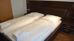 Un lit double avec du linge de lit blanc et deux oreillers, une tête de lit en bois, une table de chevet avec des tiroirs et un plancher en bois.