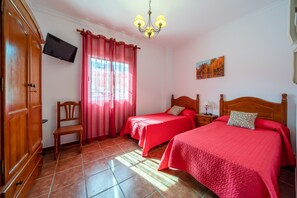 A bedroom with two beds, a TV mounted on the wall, a wooden wardrobe, a chair, and a window with curtains. - Hostal Vista a la Sierra (Valle de Abdalagis)