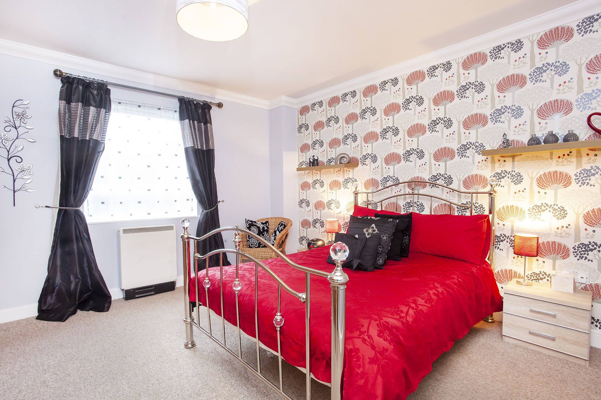 A bedroom with a metal bed frame, red bedding, a window with black curtains, a wall with tree patterns, and a bedside table with a lamp.