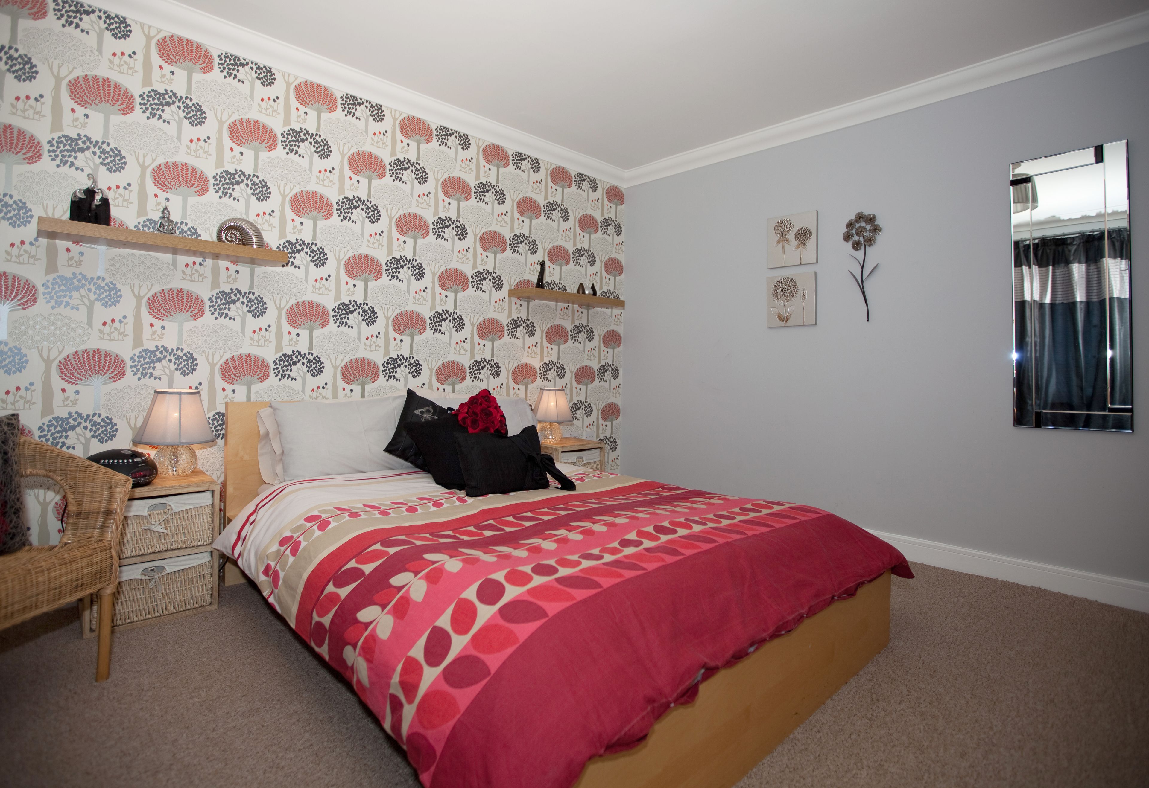 A bedroom with a patterned wallpaper, a bed with a red and white bedspread, a nightstand with a lamp, and a mirror on the wall.
