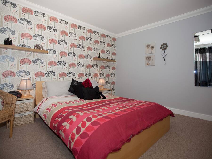 Una camera da letto con carta da parati decorata, un letto con copriletto rosso e bianco, un comodino con una lampada e uno specchio a parete.