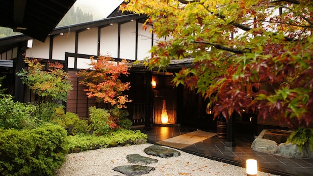 Een traditioneel Japans gebouw met een betegelde gevel, een pad van stenen en verschillende planten en bomen.