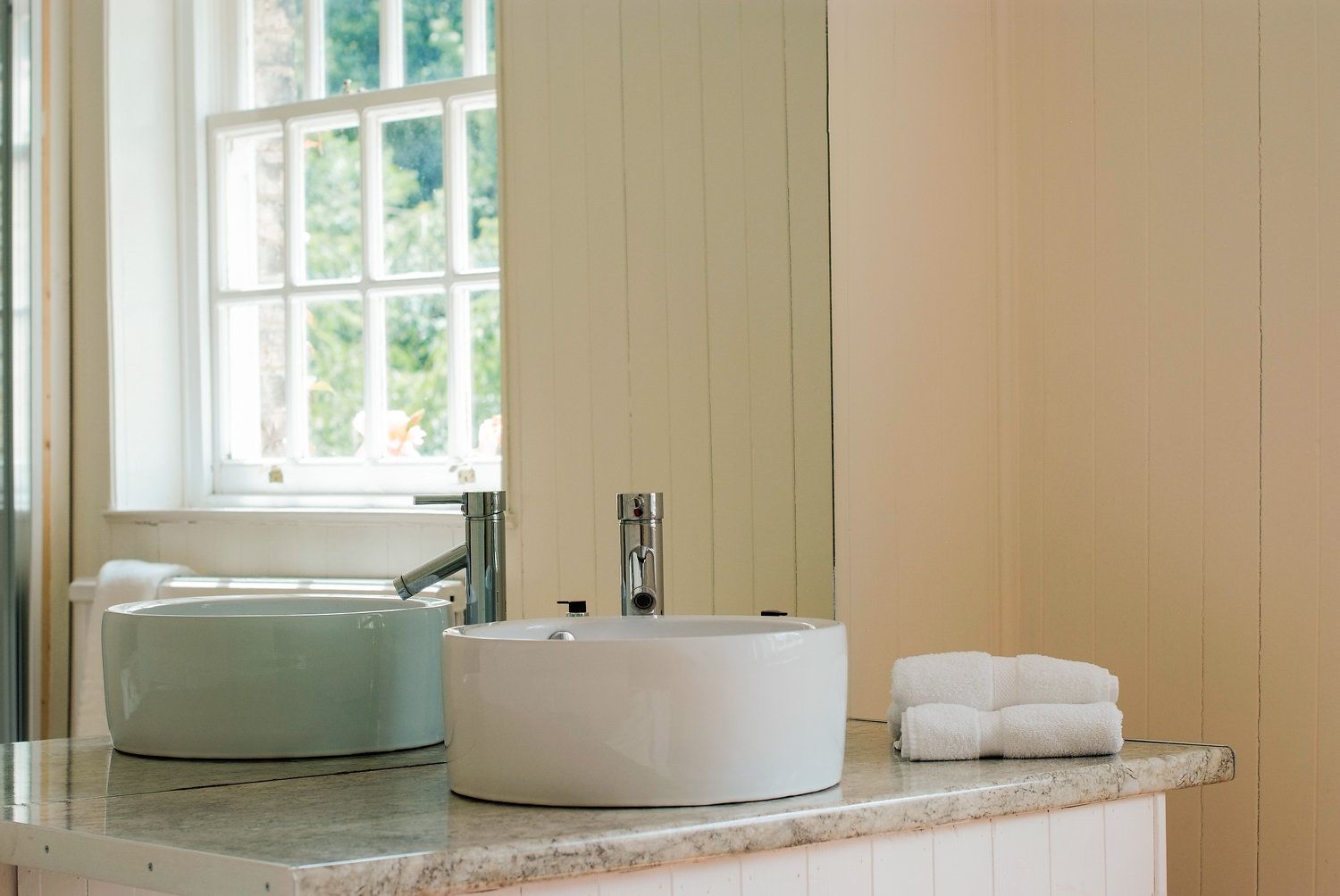 Un baño con un lavabo blanco redondo, una pileta blanca rectangular y una ventana con vista a follaje.