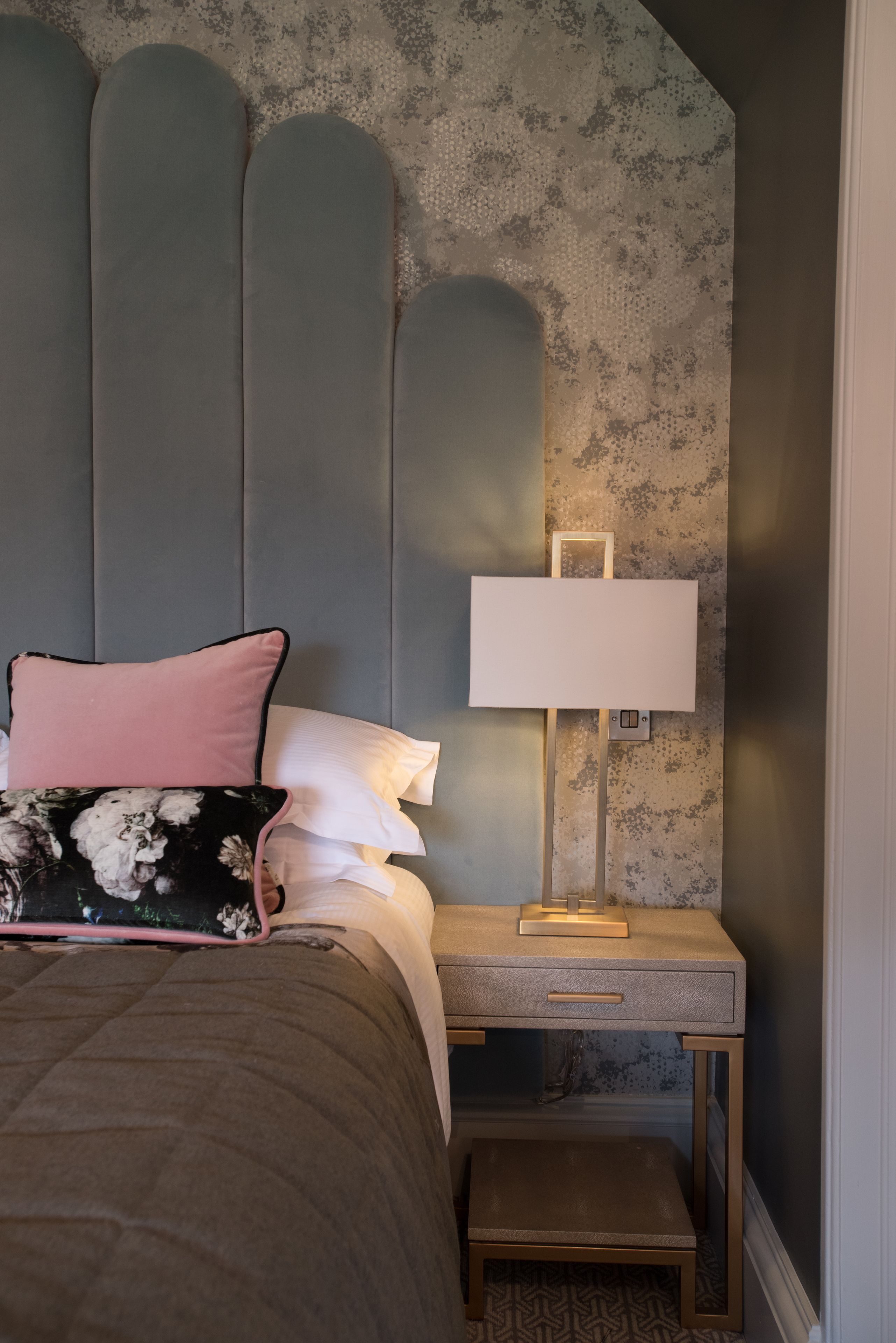 Een slaapkamer met een bed, kussens, een nachtkastje met een lamp, en een klein bankje.