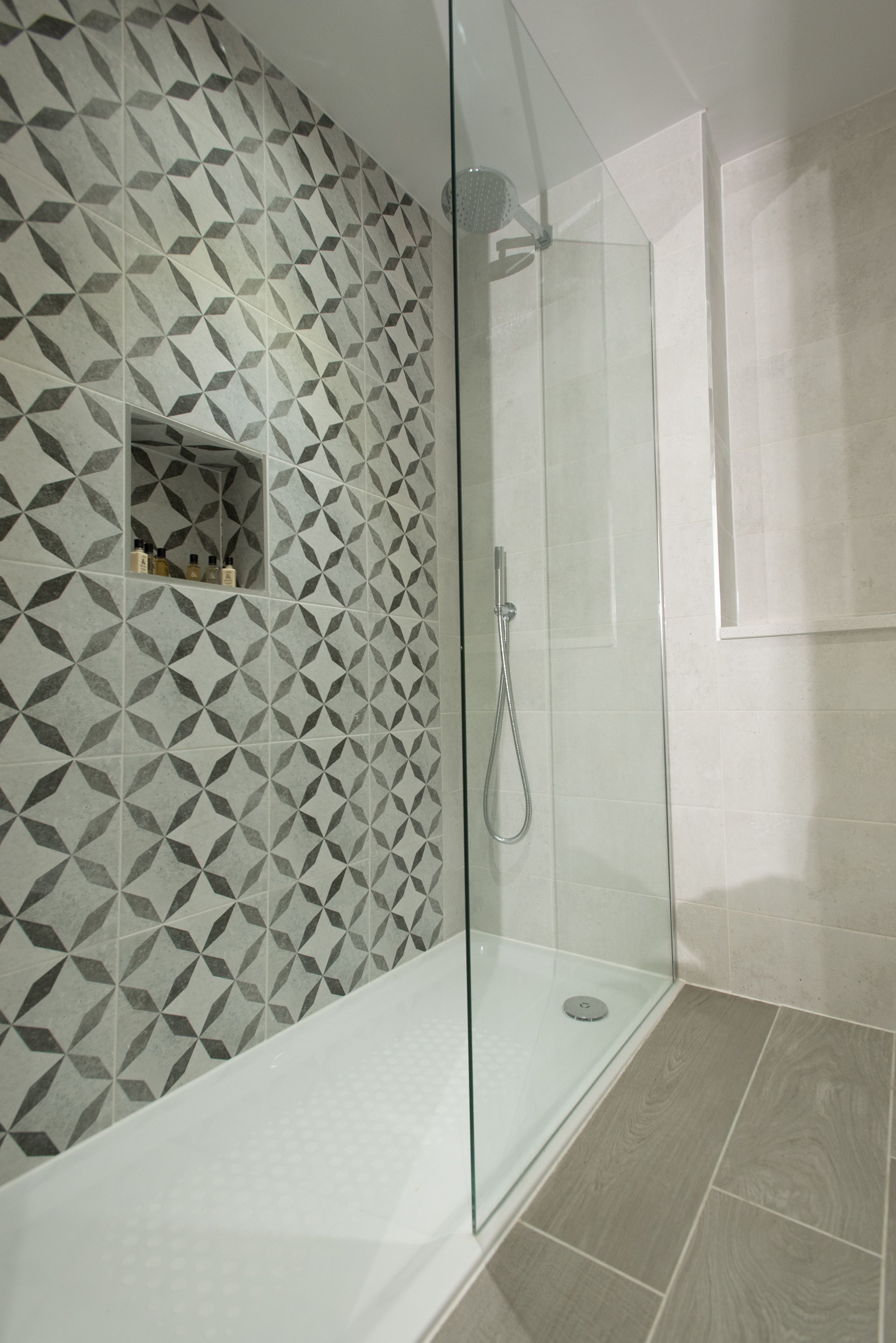 Ducha con paredes de vidrio y un patrón geométrico de azulejos en la pared, además de un estante de madera con artículos de aseo.