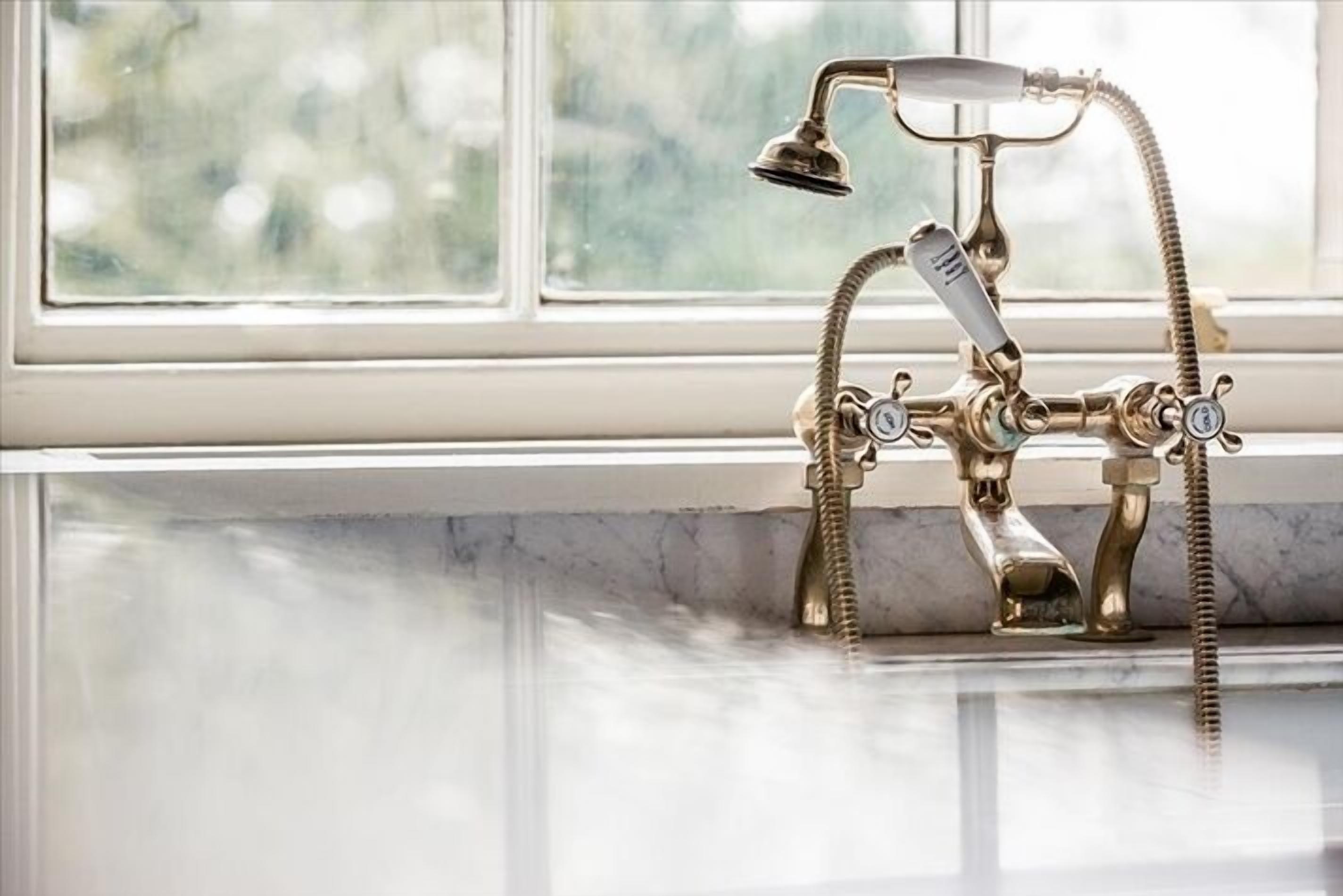 A vintage brass bathroom fixture with a shower head and multiple handles, installed in front of a window with a view of greenery.