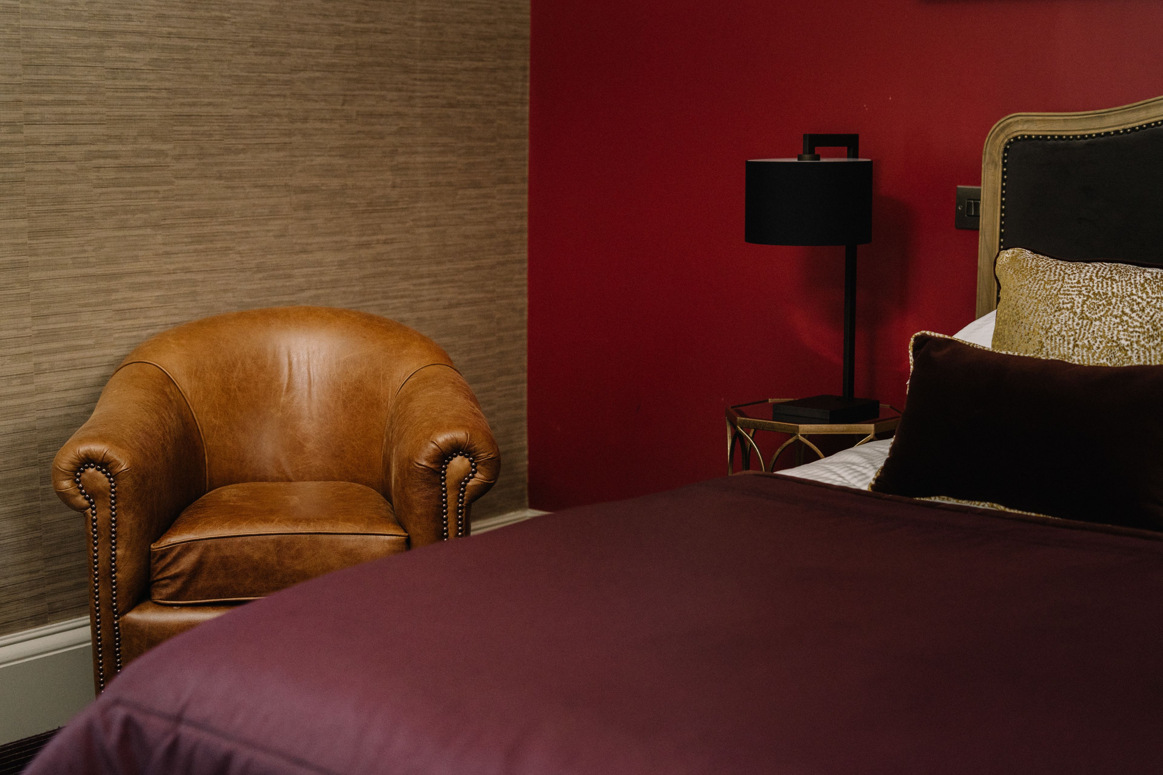 Una habitación con un sillón de cuero marrón, una cama con pared roja y una mesita de noche con una lámpara.