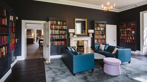 A spacious living room with dark walls, a fireplace, and multiple bookshelves filled with books. There are two blue sofas and a pink ottoman. - Saltmarshe Hall (Goole)