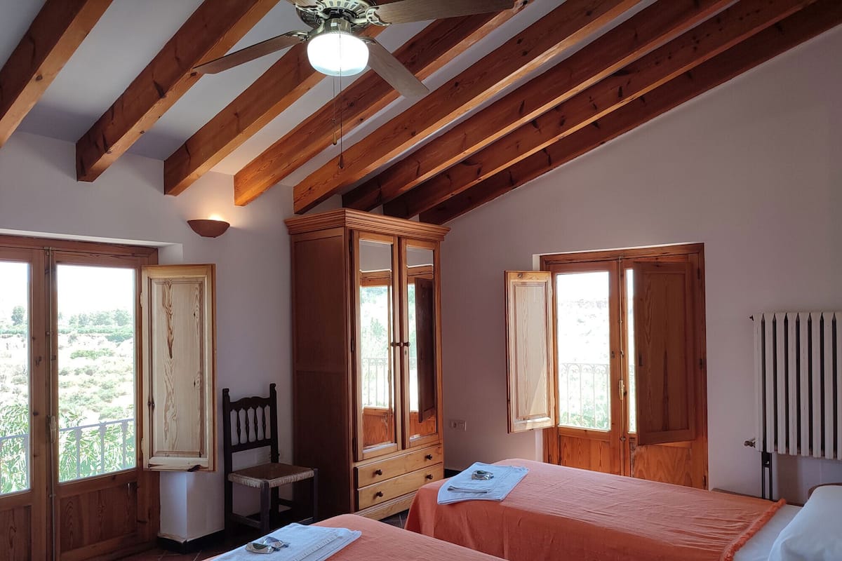 Ein Schlafzimmer mit zwei Betten, Holzbalken, einem Deckenventilator und großen Fenstern.