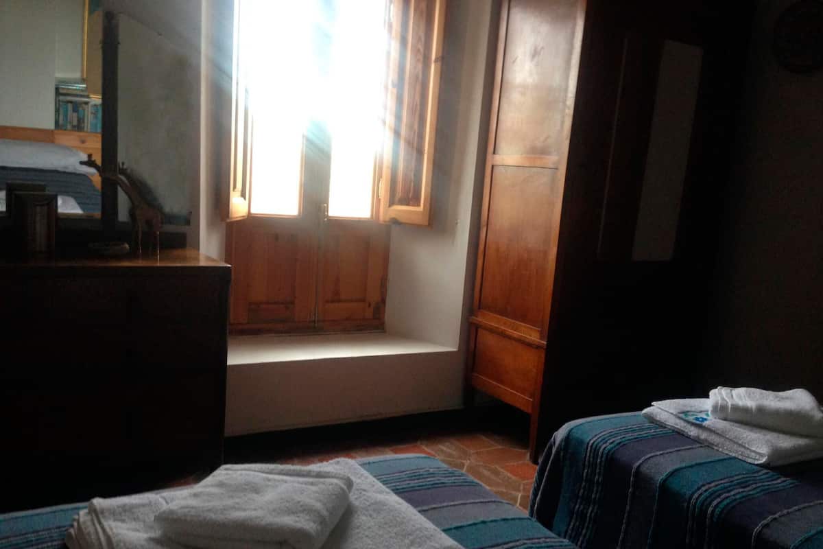 Ein kleiner, schummrig beleuchteter Raum mit einer hölzernen Bank, einem Bett mit Streifenmuster und einem Fenster mit Holzklappläden.