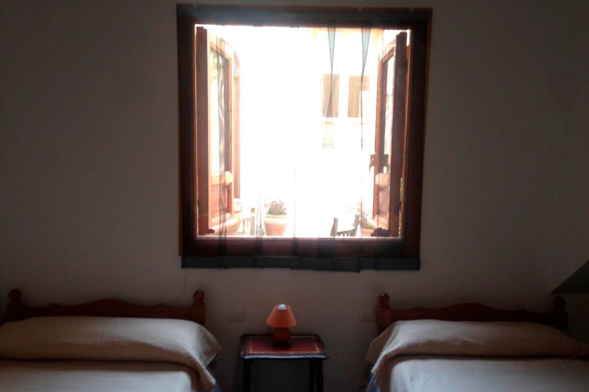 Ein Zimmer mit zwei Betten, einem kleinen Tisch mit Lampe und einem Fenster mit Vorhängen.