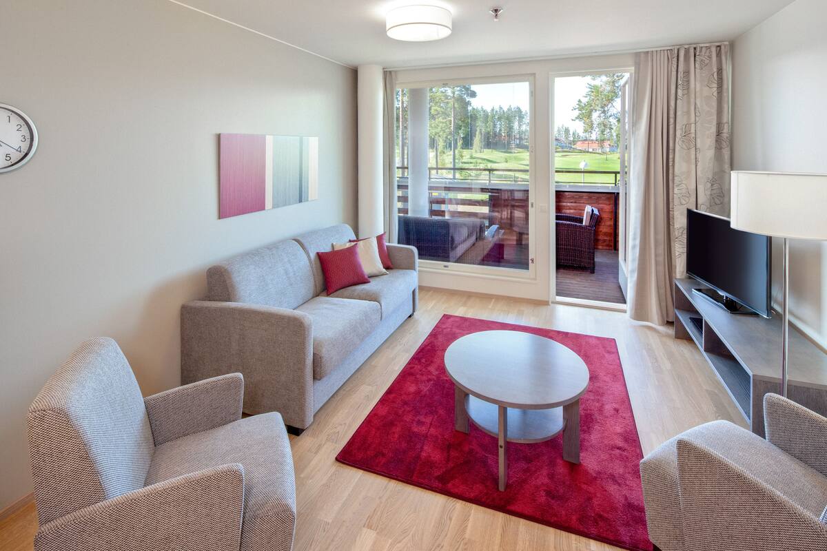 Ein modernes Wohnzimmer mit einem grauen Sofa, zwei Sesseln, einem roten Teppich, einem kleinen runden Tisch, einem Fernseher und einer Schiebetür, die auf einen Balkon mit Blick auf einen Golfplatz führt.
