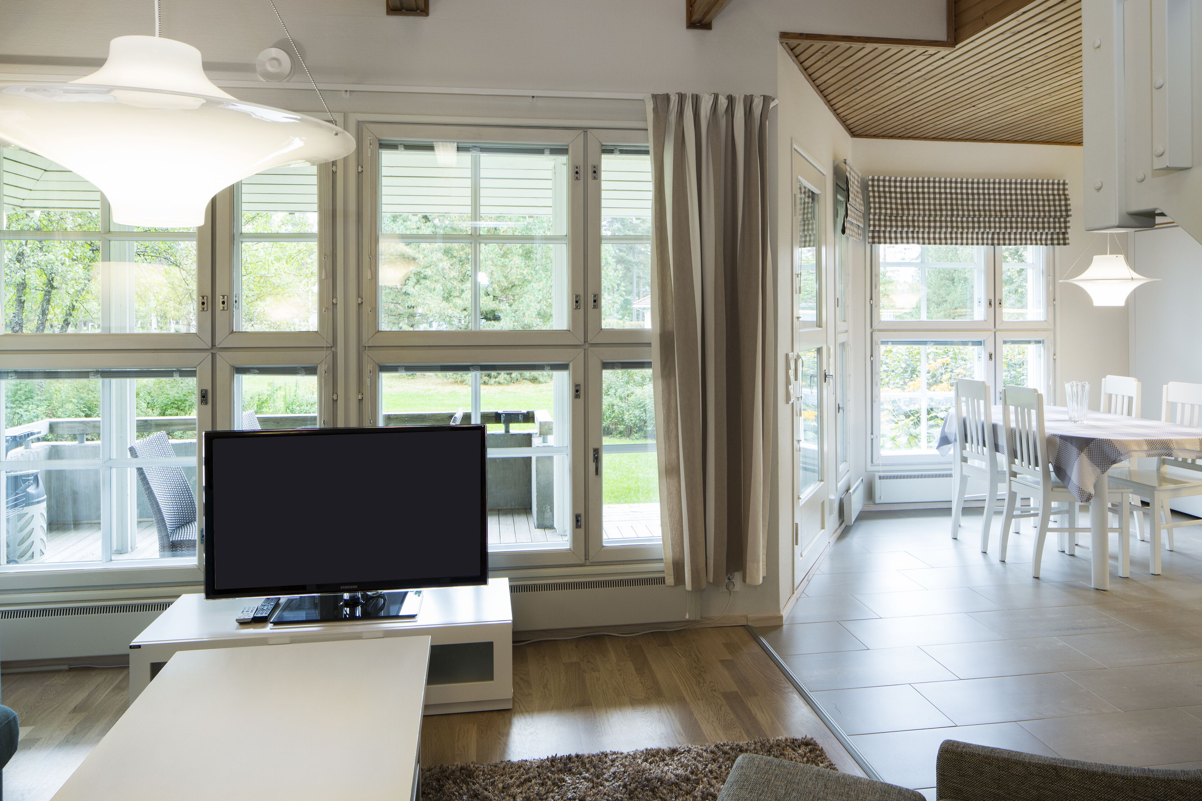 Una sala de estar moderna con un televisor de pantalla plana sobre un soporte blanco, ventanas grandes con cortinas, una zona de comedor con sillas y una mesa blancas, y un techo de madera.