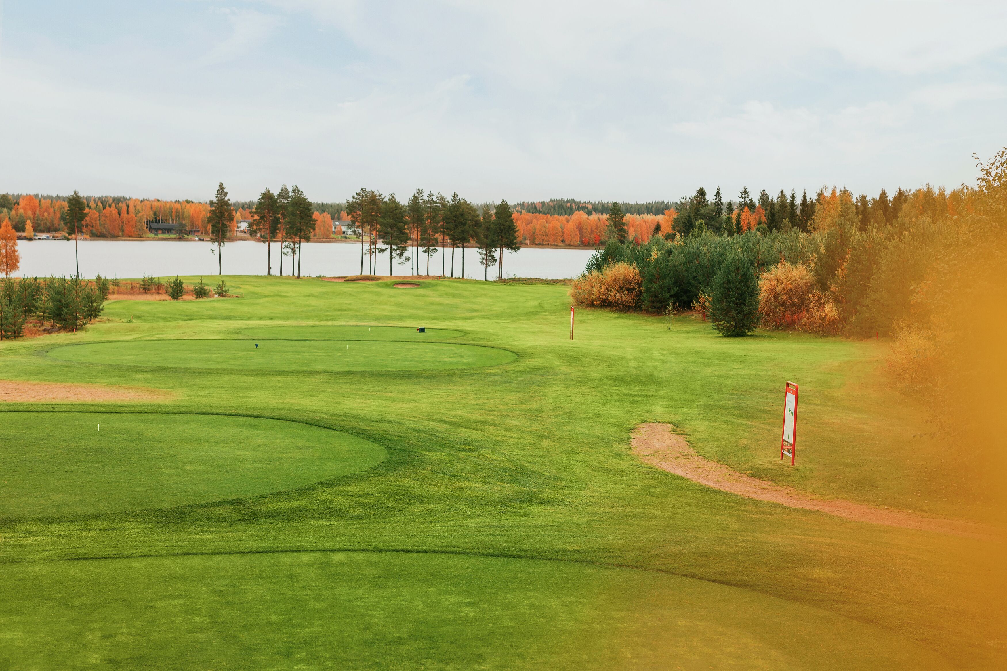 A golf course with a water body in the background, surrounded by trees with autumn foliage.