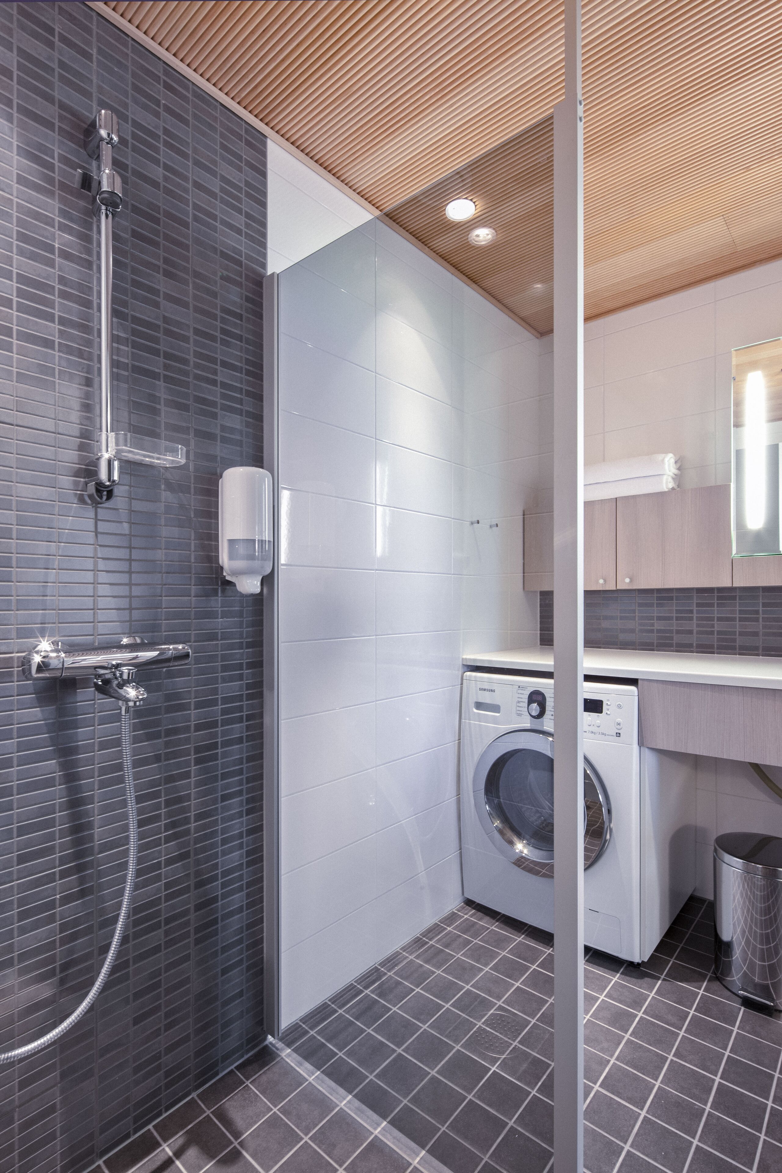 A modern bathroom with a shower, a washing machine, and a trash bin.