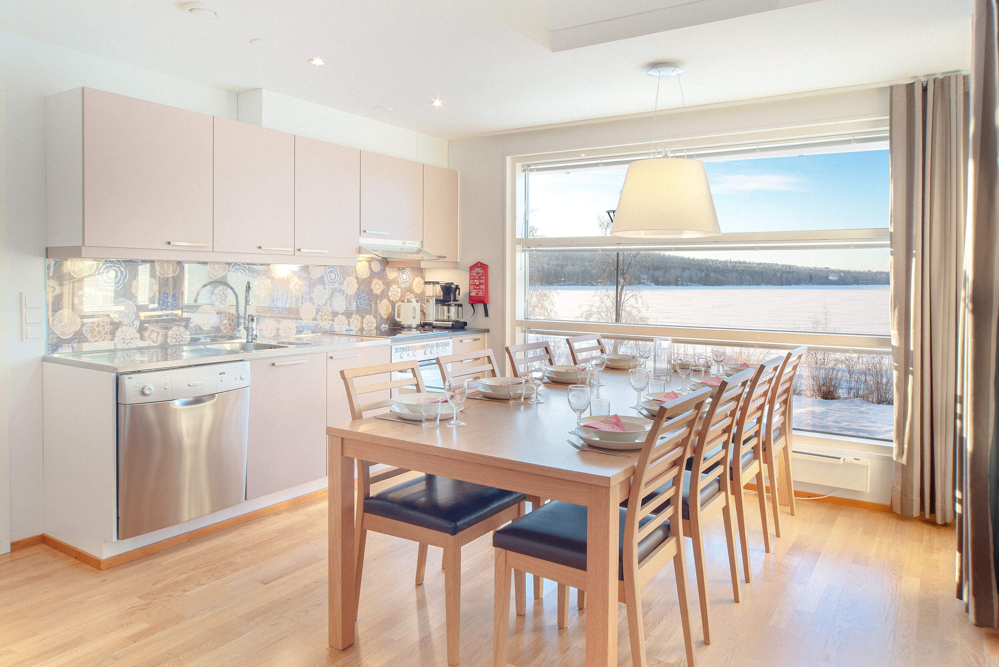 A modern kitchen with a dishwasher, a dining table set for six with chairs, and a large window overlooking a snowy landscape.