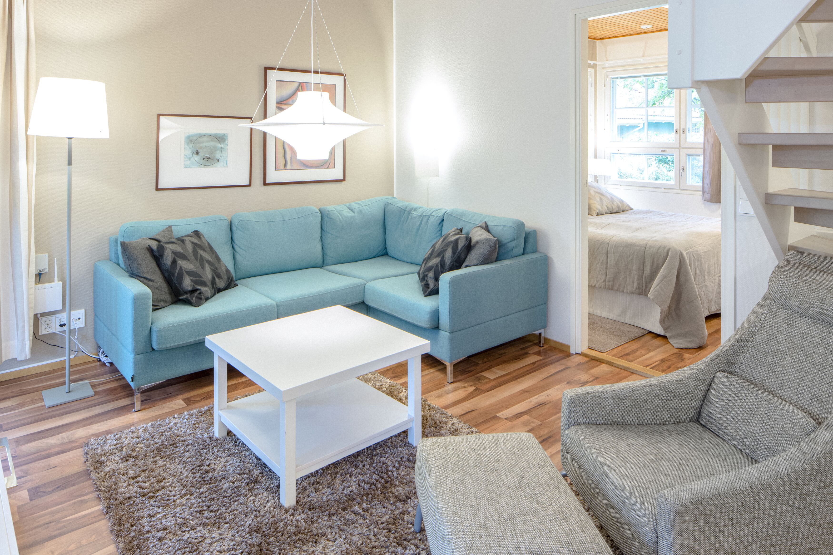 A modern living room with a light blue sectional sofa, a white coffee table, and a grey armchair. There is a large window providing natural light and a view of the outdoors.