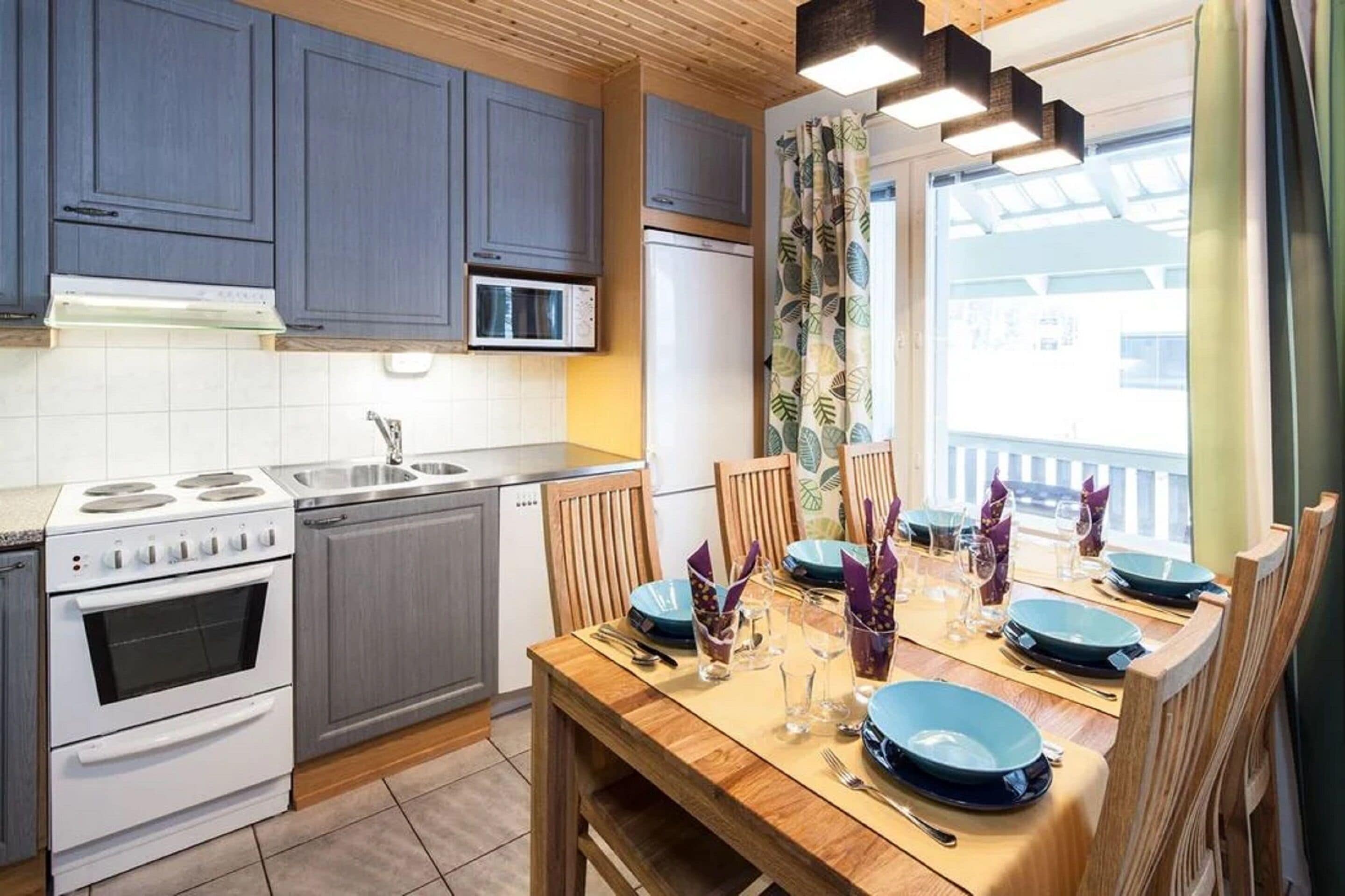 Een moderne keuken met grijze kastjes, een witte kookplaat en een eettafel voor vier personen, gedekt met blauwe borden en glazen.