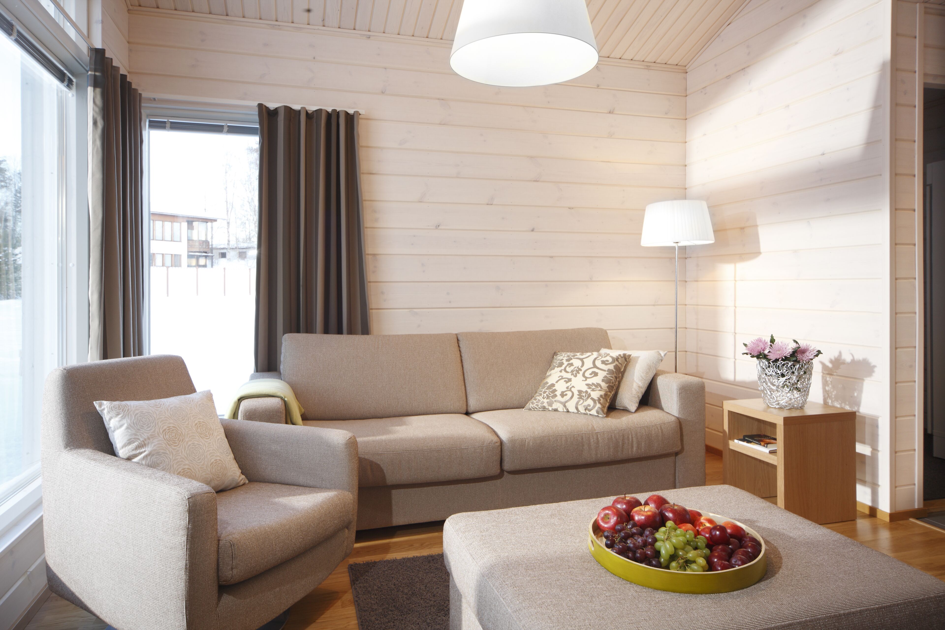 Un salon moderne avec un canapé beige, un fauteuil assorti, une table basse en bois sur laquelle se trouve un panier de fruits, et une table d’appoint avec une lampe.
