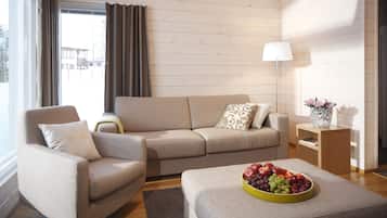 Un salon moderne avec un canapé beige, un fauteuil assorti, une table basse en bois sur laquelle se trouve un panier de fruits, et une table d’appoint avec une lampe.