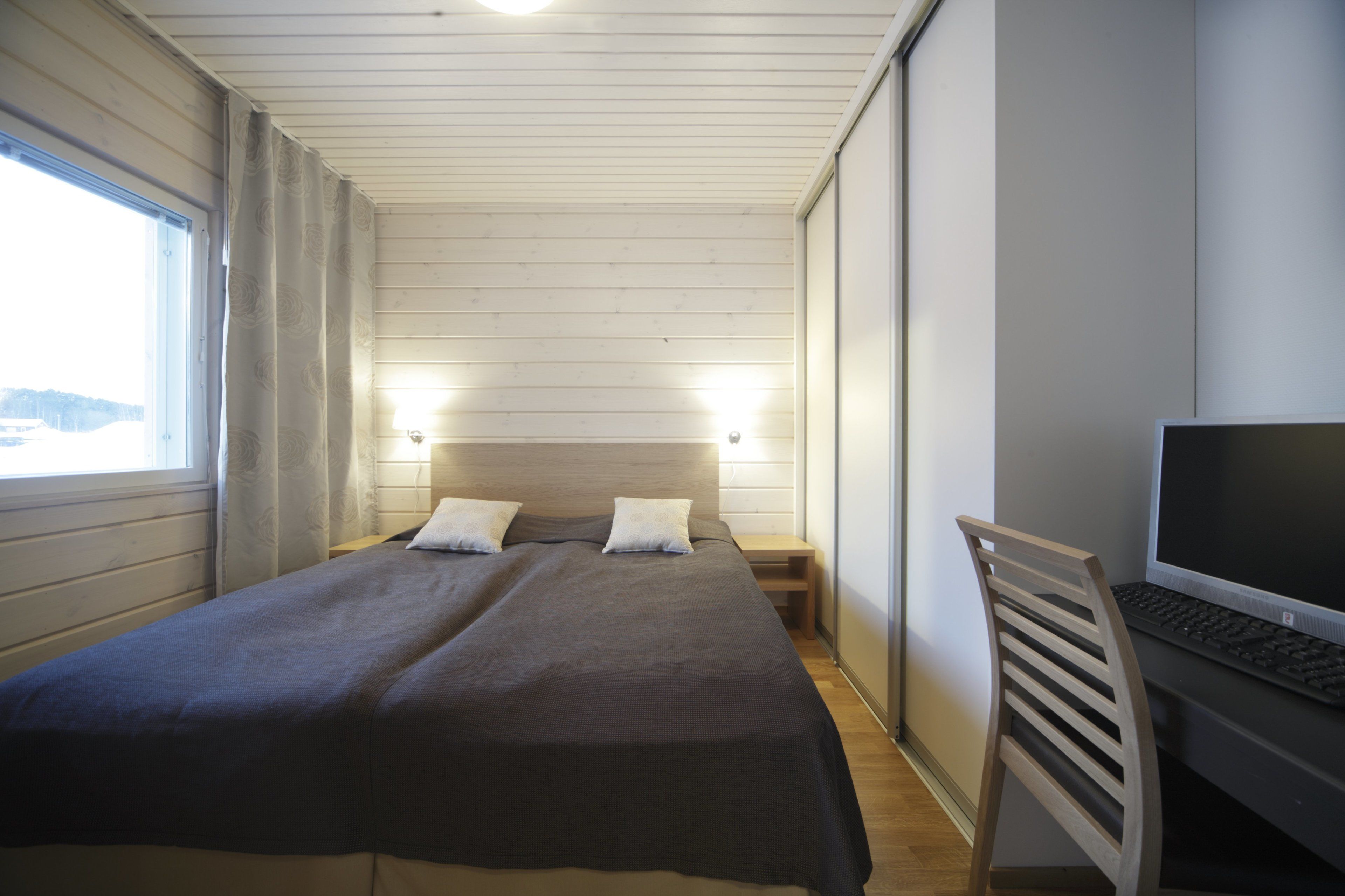 A bedroom with a bed, bedside tables, a desk with a computer, and a window.